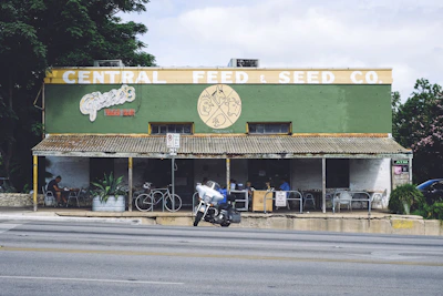 A vintage-style building with a sign reading 'Central Feed & Seed Co.' The structure has a green and yellow facade with a rustic, corrugated metal roof. Below, there is a taco bar named 'Güero's,' with outdoor seating where a few people are dining. A couple of bicycles are parked outside. A motorcycle is parked on the road in front of the building. Lush greenery surrounds the area, and there is an ATM machine visible on the right.