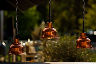 Three copper-colored hanging pendant lights are suspended in an outdoor setting with a blurred background showcasing greenery. The lights have a shiny and reflective surface, creating an elegant and warm atmosphere.
