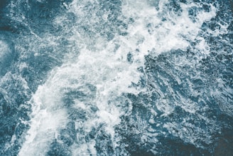 A dynamic top-down view of turbulent water, capturing the movement and texture of foamy waves and dark, deep water.