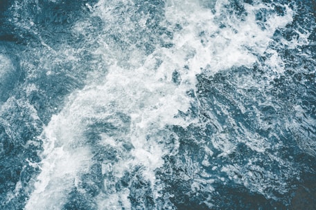 A dynamic top-down view of turbulent water, capturing the movement and texture of foamy waves and dark, deep water.