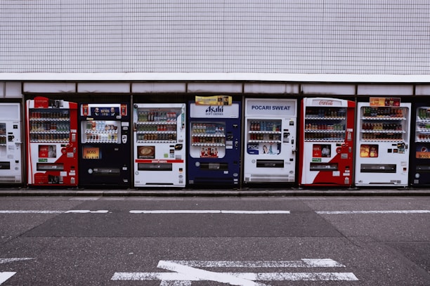 A row of vending machines is lined up on a sidewalk against a tiled wall. Each machine displays various drinks and snacks with colorful branding, including well-known beverages like Coca-Cola and Pocari Sweat. The setting appears urban, with typical street markings visible on the asphalt in front.