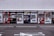A row of vending machines is lined up on a sidewalk against a tiled wall. Each machine displays various drinks and snacks with colorful branding, including well-known beverages like Coca-Cola and Pocari Sweat. The setting appears urban, with typical street markings visible on the asphalt in front.