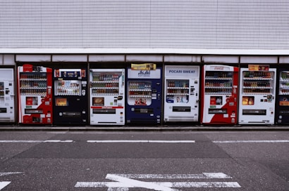 A row of vending machines is lined up on a sidewalk against a tiled wall. Each machine displays various drinks and snacks with colorful branding, including well-known beverages like Coca-Cola and Pocari Sweat. The setting appears urban, with typical street markings visible on the asphalt in front.