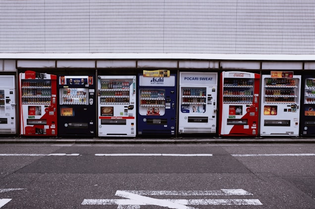 A row of vending machines is lined up on a sidewalk against a tiled wall. Each machine displays various drinks and snacks with colorful branding, including well-known beverages like Coca-Cola and Pocari Sweat. The setting appears urban, with typical street markings visible on the asphalt in front.