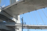 A large bridge made of concrete and steel cables spans overhead. The structure is supported by thick vertical pillars, and numerous cables stretch diagonally, creating a geometric pattern against the clear blue sky. Below the bridge, power lines run vertically, adding complexity to the scene.