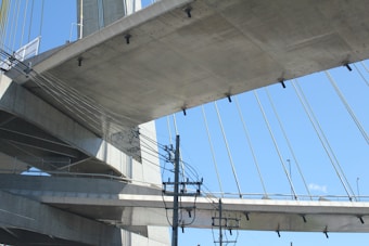 A large bridge made of concrete and steel cables spans overhead. The structure is supported by thick vertical pillars, and numerous cables stretch diagonally, creating a geometric pattern against the clear blue sky. Below the bridge, power lines run vertically, adding complexity to the scene.