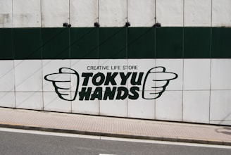 A large sign on a wall featuring the text 'TOKYU HANDS' with the phrase 'CREATIVE LIFE STORE' above it. The logo includes the outline of two hands, one on each side of the text. The wall is divided into green and white sections, and there is a sidewalk and street in front of it.
