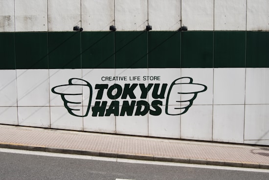 A large sign on a wall featuring the text 'TOKYU HANDS' with the phrase 'CREATIVE LIFE STORE' above it. The logo includes the outline of two hands, one on each side of the text. The wall is divided into green and white sections, and there is a sidewalk and street in front of it.