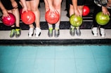 Five people standing side by side are holding colorful bowling balls in their hands. They are wearing bowling shoes with various designs, and their feet are positioned on a tiled floor with a mixture of blue and black tiles.