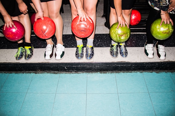 Five people standing side by side are holding colorful bowling balls in their hands. They are wearing bowling shoes with various designs, and their feet are positioned on a tiled floor with a mixture of blue and black tiles.
