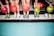 Five people standing side by side are holding colorful bowling balls in their hands. They are wearing bowling shoes with various designs, and their feet are positioned on a tiled floor with a mixture of blue and black tiles.