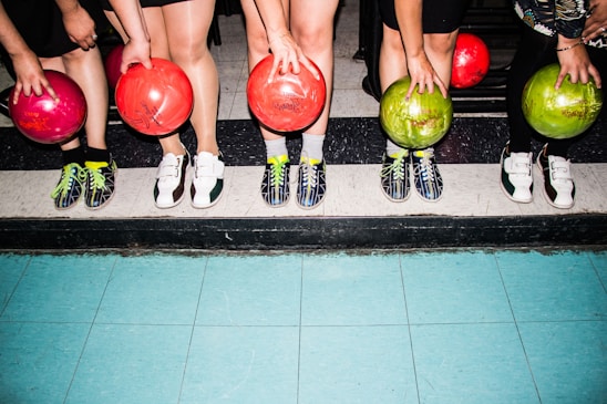 Five people standing side by side are holding colorful bowling balls in their hands. They are wearing bowling shoes with various designs, and their feet are positioned on a tiled floor with a mixture of blue and black tiles.