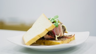A sandwich featuring two slices of white bread encasing layers of cooked bacon, ground beef, mayonnaise, and fresh green basil leaves. The sandwich is presented on a white plate set against a simple, neutral background.