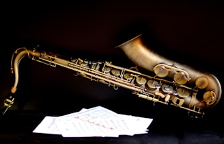 A brass saxophone is positioned horizontally against a dark background, with detailed engravings and intricate keys visible. In the foreground, sheets of music are scattered, contrasting with the black surface.
