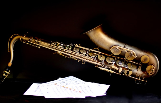 A brass saxophone is positioned horizontally against a dark background, with detailed engravings and intricate keys visible. In the foreground, sheets of music are scattered, contrasting with the black surface.