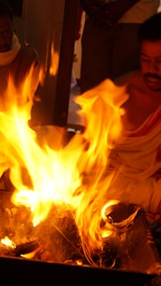 Vivid flames erupt from a fire pit, surrounded by individuals engaged in a ritual. They wear traditional attire and seem to be participating in a spiritual or religious ceremony.