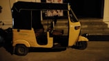 A yellow auto rickshaw is parked on a dimly lit street next to a building with steps leading to an entrance. The vehicle has a black roof and three wheels, with some text visible on its side.