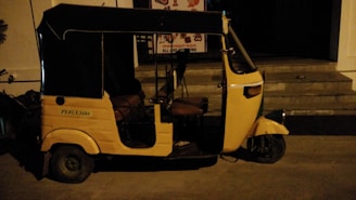 A yellow auto rickshaw is parked on a dimly lit street next to a building with steps leading to an entrance. The vehicle has a black roof and three wheels, with some text visible on its side.