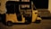 A yellow auto rickshaw is parked on a dimly lit street next to a building with steps leading to an entrance. The vehicle has a black roof and three wheels, with some text visible on its side.