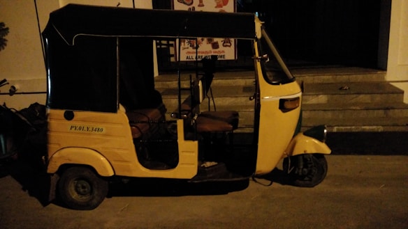 A yellow auto rickshaw is parked on a dimly lit street next to a building with steps leading to an entrance. The vehicle has a black roof and three wheels, with some text visible on its side.