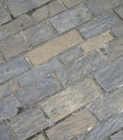 Gray and beige stone tiles laid out in a rectangular brick pattern. The surface appears weathered with some small cracks and variations in texture and color, giving it a rugged and natural appearance.
