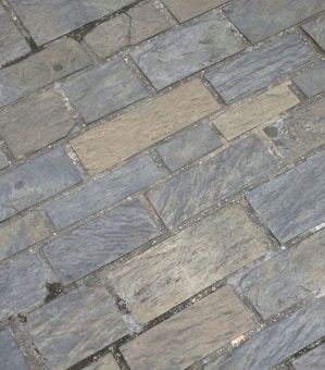 Gray and beige stone tiles laid out in a rectangular brick pattern. The surface appears weathered with some small cracks and variations in texture and color, giving it a rugged and natural appearance.