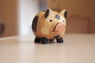 A ceramic or wooden piggy bank sits on a light wooden table. The piggy bank is painted with beige, black, and pink colors, showcasing a stylized and simplistic design. Its eyes are small and red, adding a playful element to its appearance.