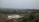 A landscape featuring a white building under construction situated on a red dirt plot, surrounded by lush green fields and rolling hills. In the distance, patches of greenery are interspersed with clusters of buildings, creating a rural setting.