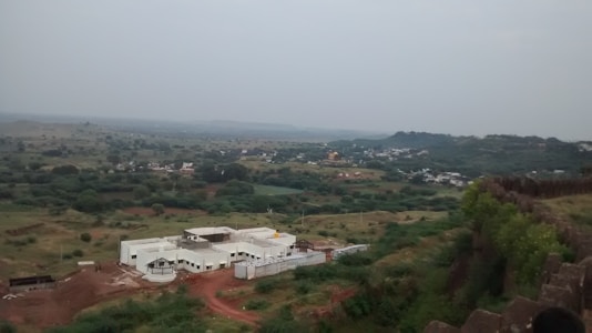 A landscape featuring a white building under construction situated on a red dirt plot, surrounded by lush green fields and rolling hills. In the distance, patches of greenery are interspersed with clusters of buildings, creating a rural setting.