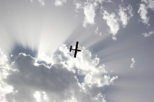 A silhouette of a small aircraft is seen flying against a dramatic sky, with beams of sunlight streaming through scattered clouds.