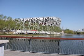 A large, modern stadium with a distinctive white lattice structure stands beside a body of water. The structure is surrounded by trees, and a railing occupies the foreground. Clear blue skies and a few cranes and buildings can be seen in the background.