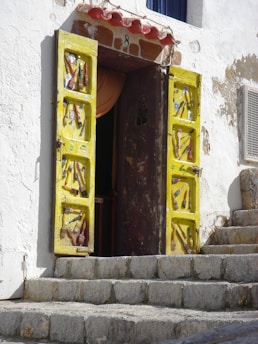 A rustic doorway with bright yellow doors adorned with various stickers and decorations. The doorway is set in a textured white wall with visible signs of wear and age. Stone steps lead up to the entrance, enhancing the building's vintage charm. Above the door, there is a red-tiled canopy and the number 8 is displayed.