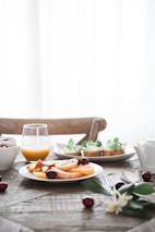 A rustic wooden table set with a variety of breakfast items, including a plate of sliced fruits garnished with cherries, a glass of orange juice, a small teapot, and other dishes featuring herbs like mint and basil. The background is softly lit with natural light coming through sheer curtains.