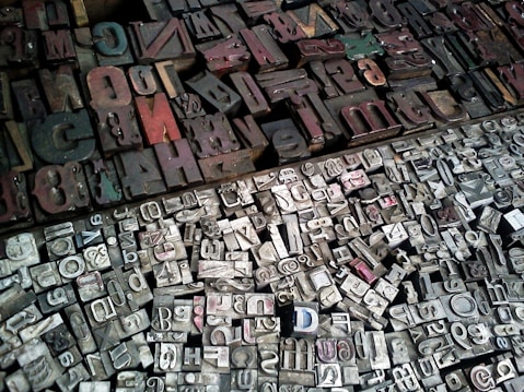 A diverse collection of vintage letterpress printing blocks arranged closely together. The blocks vary in size, shape, and color, featuring a mix of uppercase and lowercase letters, numbers, and symbols. The surface is mostly metallic with signs of wear and age, reflecting a historical or industrial atmosphere.