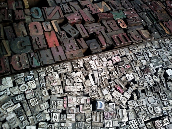 A diverse collection of vintage letterpress printing blocks arranged closely together. The blocks vary in size, shape, and color, featuring a mix of uppercase and lowercase letters, numbers, and symbols. The surface is mostly metallic with signs of wear and age, reflecting a historical or industrial atmosphere.