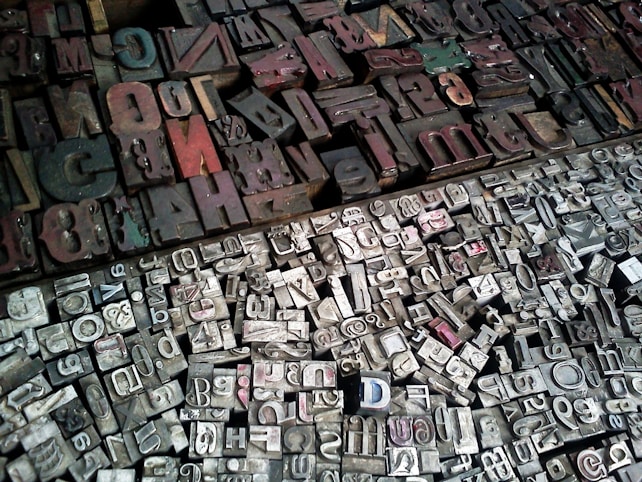 A diverse collection of vintage letterpress printing blocks arranged closely together. The blocks vary in size, shape, and color, featuring a mix of uppercase and lowercase letters, numbers, and symbols. The surface is mostly metallic with signs of wear and age, reflecting a historical or industrial atmosphere.