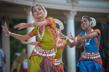 A group of dancers wearing traditional Indian attire adorned with ornate jewelry perform expressive dance movements. The colorful garments include a mix of bright green, blue, and red fabrics, intricately decorated with silver embellishments. The dancers hold graceful poses, demonstrating elegance and skill.