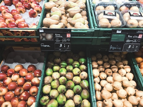 An assortment of fruits is displayed in green plastic containers at a market or grocery store. The selection includes red apples, green pears, and brownish pears placed alongside packages of mushrooms. Price tags with country origins such as France and Argentina are visible above the fruits.