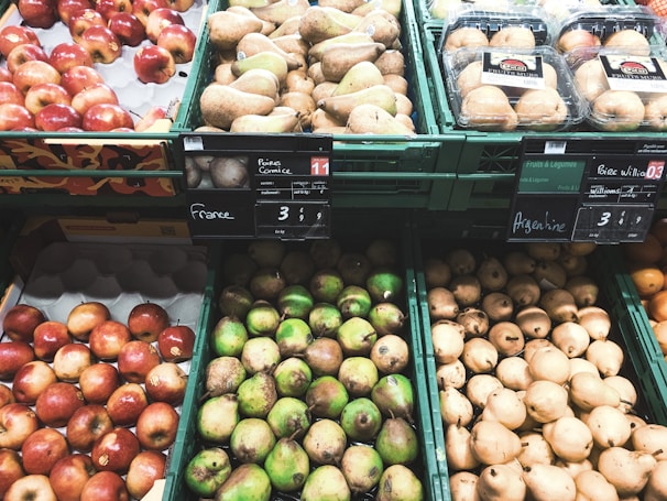 An assortment of fruits is displayed in green plastic containers at a market or grocery store. The selection includes red apples, green pears, and brownish pears placed alongside packages of mushrooms. Price tags with country origins such as France and Argentina are visible above the fruits.