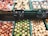 An assortment of fruits is displayed in green plastic containers at a market or grocery store. The selection includes red apples, green pears, and brownish pears placed alongside packages of mushrooms. Price tags with country origins such as France and Argentina are visible above the fruits.