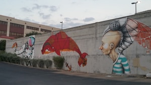 A large mural on a concrete wall depicting a colorful astronaut reaching out towards a slice of pizza, a red dinosaur-like creature, and a person with an insect-like mechanical head wearing a striped shirt. The background features a building with a red roof and windows.