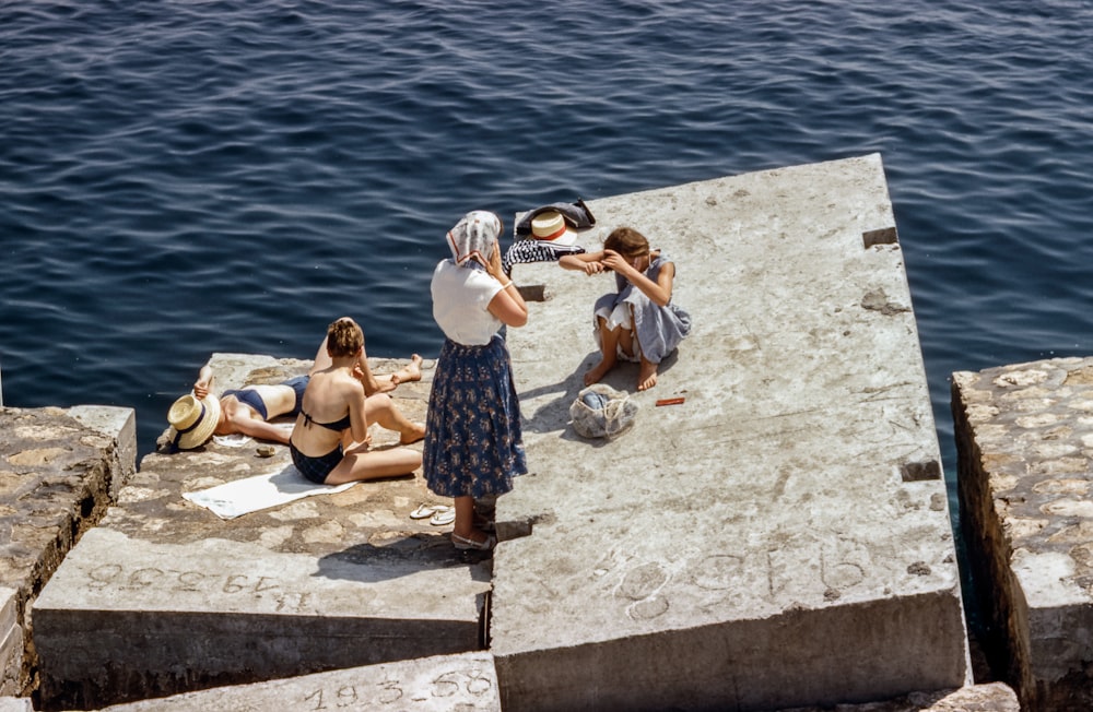 Frauen in Badeanzügen und Vintage-Kleidung sonnen sich auf Zementwürfeln am Meer