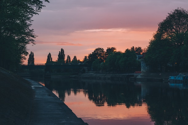 A serene riverside scene at sunset with a wooden footbridge and lush tropical forest.