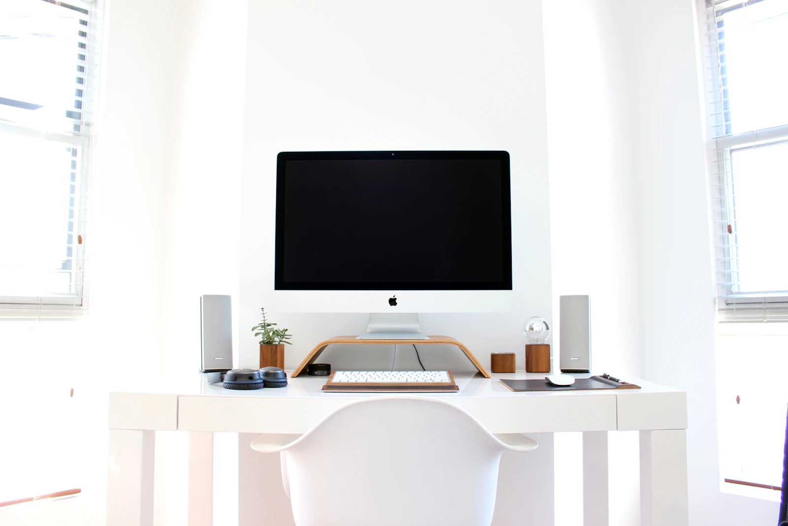 All-white minimalist home office with a desktop computer, small speakers and a potted plant on a white desk between two bright windows