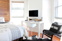 A modern, minimalist bedroom with a focus on clean lines and simplicity. The room features a bed with a geometric patterned duvet and a white chair at a sleek white desk holding a computer. Wood panel accents on one wall add warmth, with large windows allowing ample natural light. A decorative pillow with 'New York' is placed on a black chair, and a fur rug adds a touch of coziness.