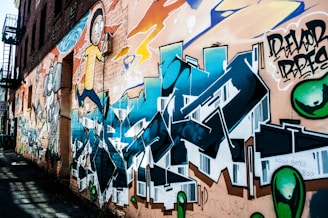 A colorful graffiti mural covers the side of an urban brick building. The artwork includes cartoon-style characters and stylized lettering in vibrant hues of blue, orange, and green. The mural features detailed shading and dynamic forms, creating a lively and energetic atmosphere.