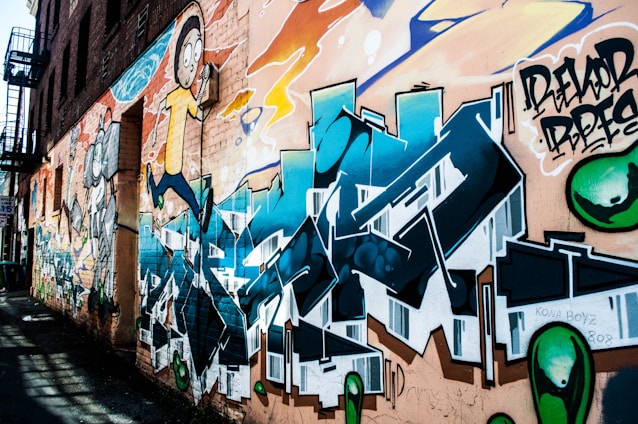 A colorful graffiti mural covers the side of an urban brick building. The artwork includes cartoon-style characters and stylized lettering in vibrant hues of blue, orange, and green. The mural features detailed shading and dynamic forms, creating a lively and energetic atmosphere.