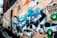 A colorful graffiti mural covers the side of an urban brick building. The artwork includes cartoon-style characters and stylized lettering in vibrant hues of blue, orange, and green. The mural features detailed shading and dynamic forms, creating a lively and energetic atmosphere.