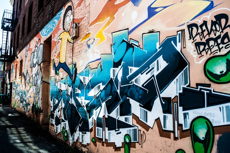 A colorful graffiti mural covers the side of an urban brick building. The artwork includes cartoon-style characters and stylized lettering in vibrant hues of blue, orange, and green. The mural features detailed shading and dynamic forms, creating a lively and energetic atmosphere.