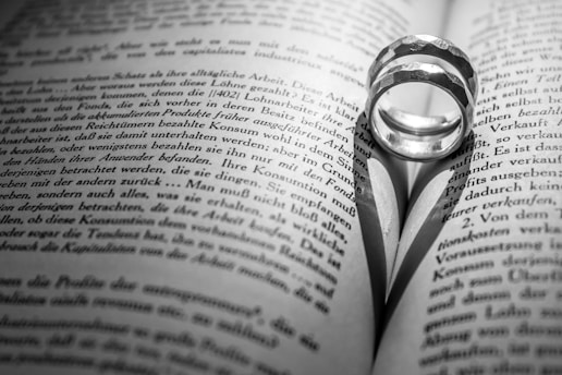 A pair of rings placed on an open book, casting a heart-shaped shadow on the pages. The text on the book is in black and white, with the focus centered on the rings and the shadow they create.